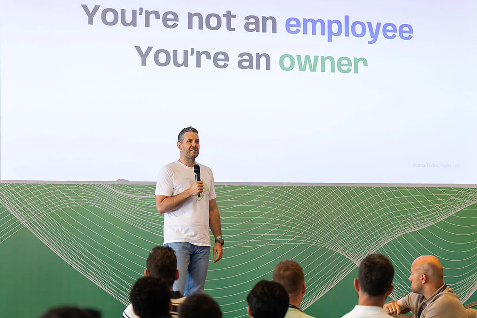 A speaker on stage beside a slide reading You're not an employee, you're an owner